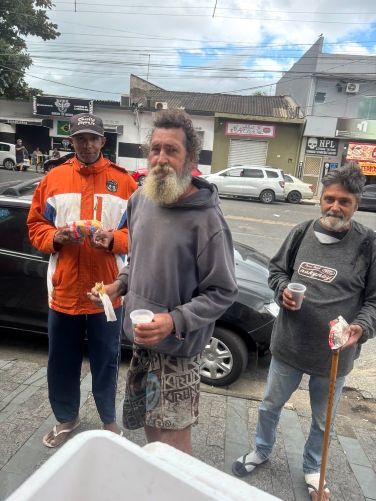 Três homens na calçada recebem lanche e um copo de café